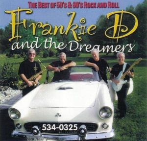 Photograph of the rock band Frankie D and the Dreamers standing with instruments and next to a white race car